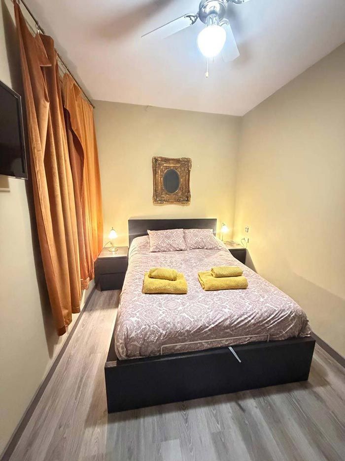Habitación pequeña con una cama, toallas amarillas dobladas, cortinas cálidas y un ventilador de techo bajo un cuadro decorativo.