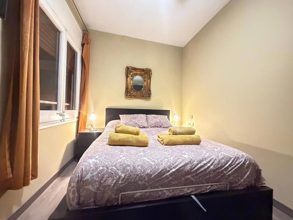 Habitación pequeña con cama hecha, toallas amarillas, lámparas de noche y una ventana con cortinas.