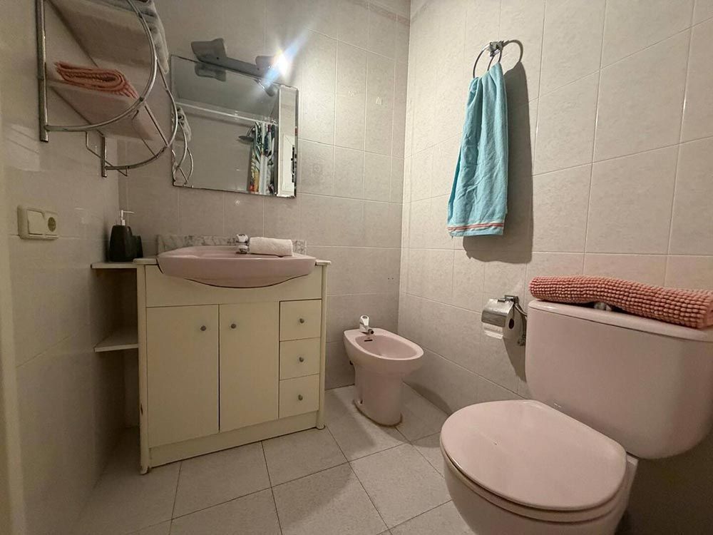 Baño pequeño de color beige con lavabo, inodoro, bidé, espejo y toalla azul sobre paredes alicatadas.