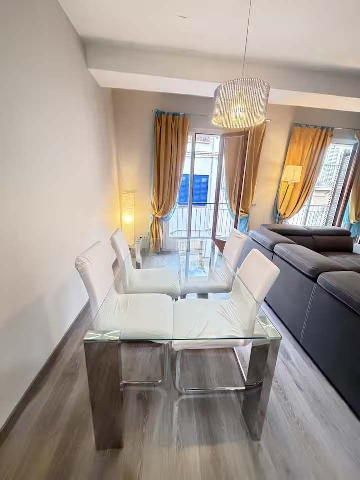 Comedor moderno con mesa de cristal, sillas blancas, lámpara colgante y sofá gris cerca de las ventanas y cortinas.