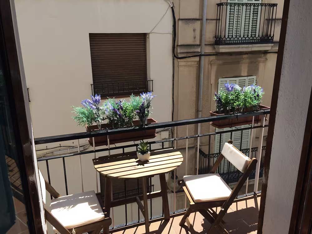 Pequeño balcón con dos sillas, una mesa redonda y jardineras con vistas a un patio interior.