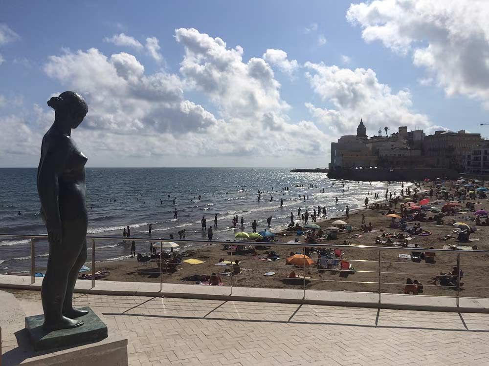 Estatua de bronce con vistas a una playa abarrotada y al mar bajo un cielo nublado.