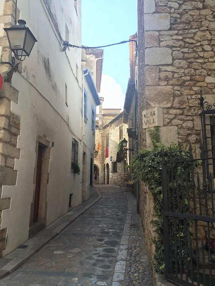Un estrecho callejón empedrado entre edificios de piedra bajo un cielo azul brillante, con farolas y barandilla de hierro.