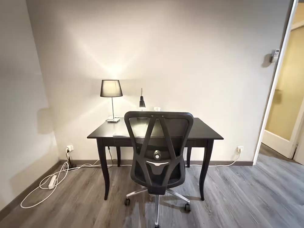Pequeña oficina en casa con escritorio negro, silla de malla, lámpara y paredes blancas sobre alfombra gris.