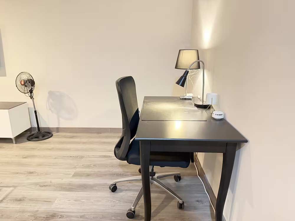 Oficina en casa minimalista con escritorio, silla, lámpara y ventilador negros contra una pared clara.