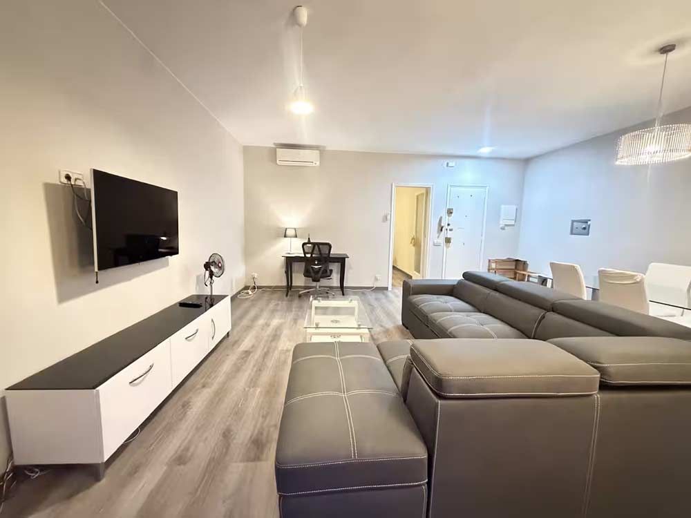 Luminoso salón moderno con sofá seccional gris, televisor montado en la pared y consola blanca en un amplio espacio diáfano.