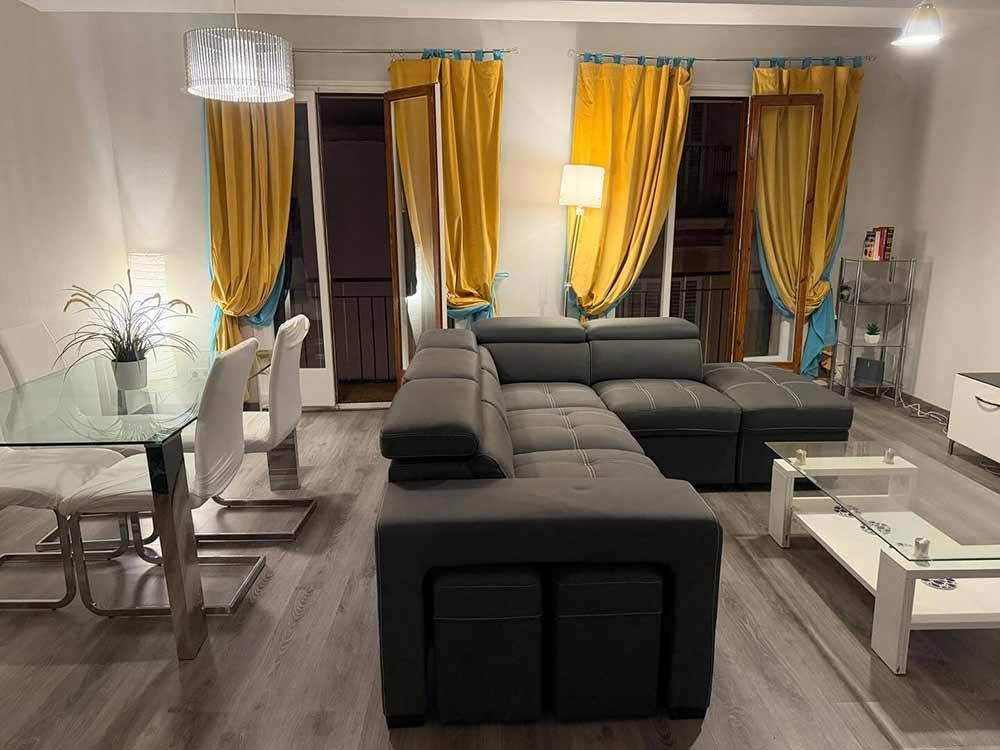 Sala de estar moderna con sofá seccional gris, mesa de comedor de cristal y cortinas amarillas junto a las ventanas.