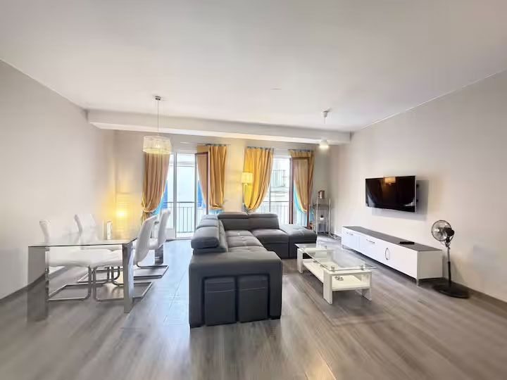 Sala de estar moderna y luminosa con sofá modular gris, muebles blancos, televisor montado en la pared y cortinas amarillas.