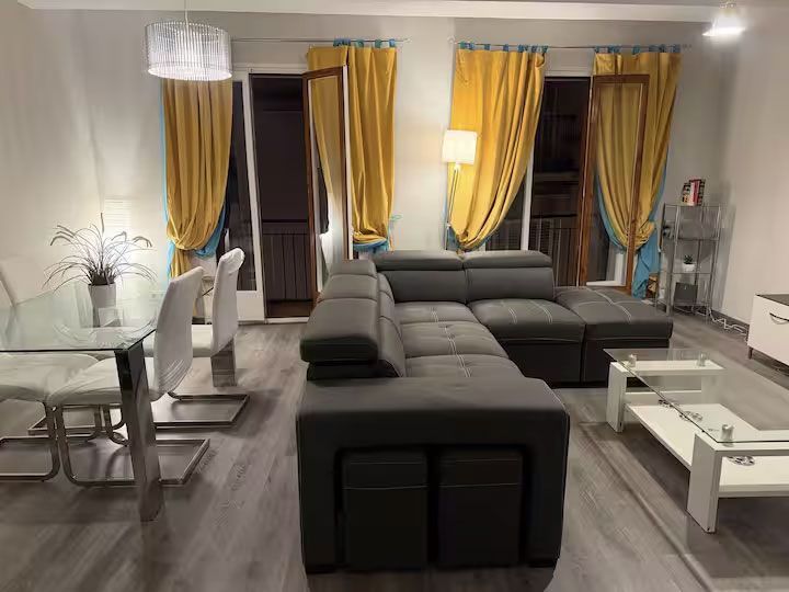 Sala de estar moderna con sofá seccional gris, mesa de centro de cristal, zona de comedor y cortinas amarillas.