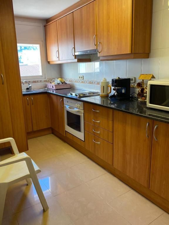 Cocina pequeña con armarios de madera, encimeras negras, horno blanco, microondas y una silla junto a la ventana.