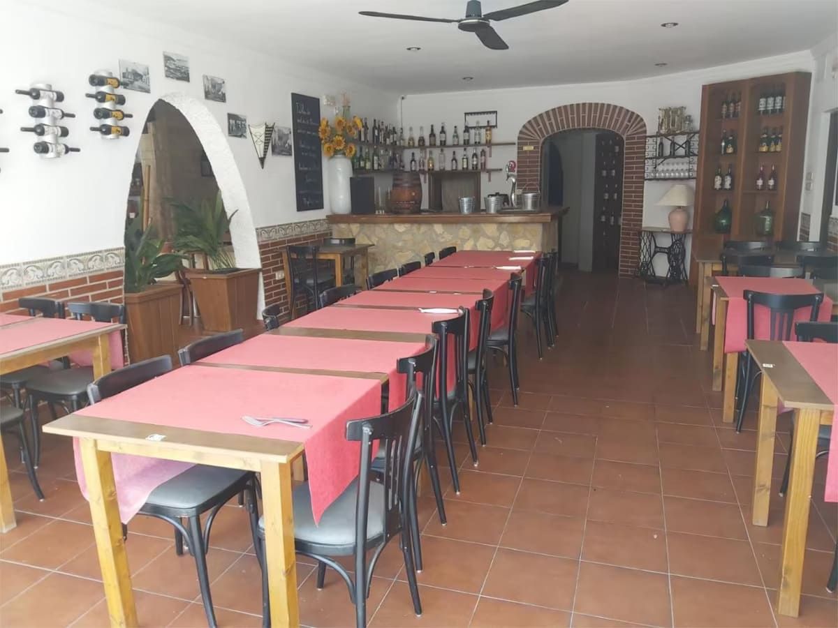 Interior de restaurante con manteles rojos, sillas negras, bar, botelleros y puertas arqueadas.