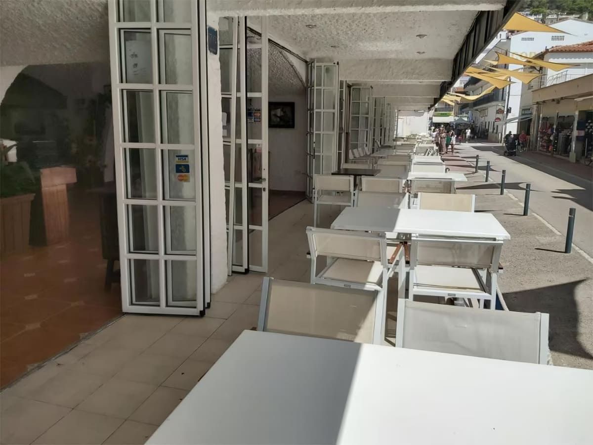 Patio de restaurante con mesas y sillas blancas, bajo una pasarela cubierta en una calle soleada.