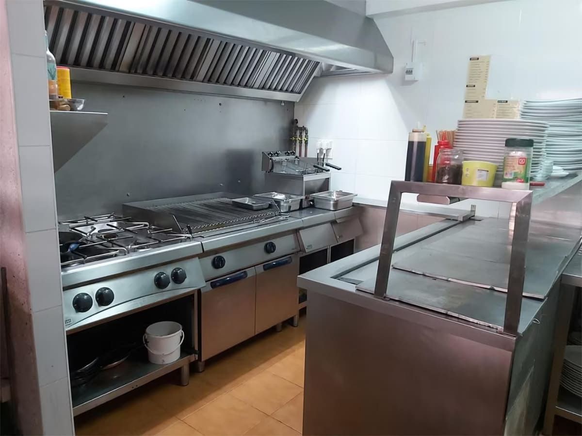 Cocina comercial con electrodomésticos de acero inoxidable, estufa, parrilla y campana extractora.