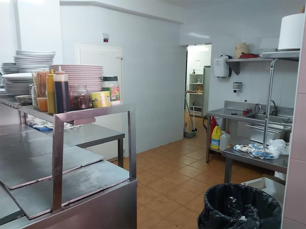 Cocina comercial con encimeras de acero inoxidable, estantes y diversos suministros. Una puerta da acceso a otra habitación.