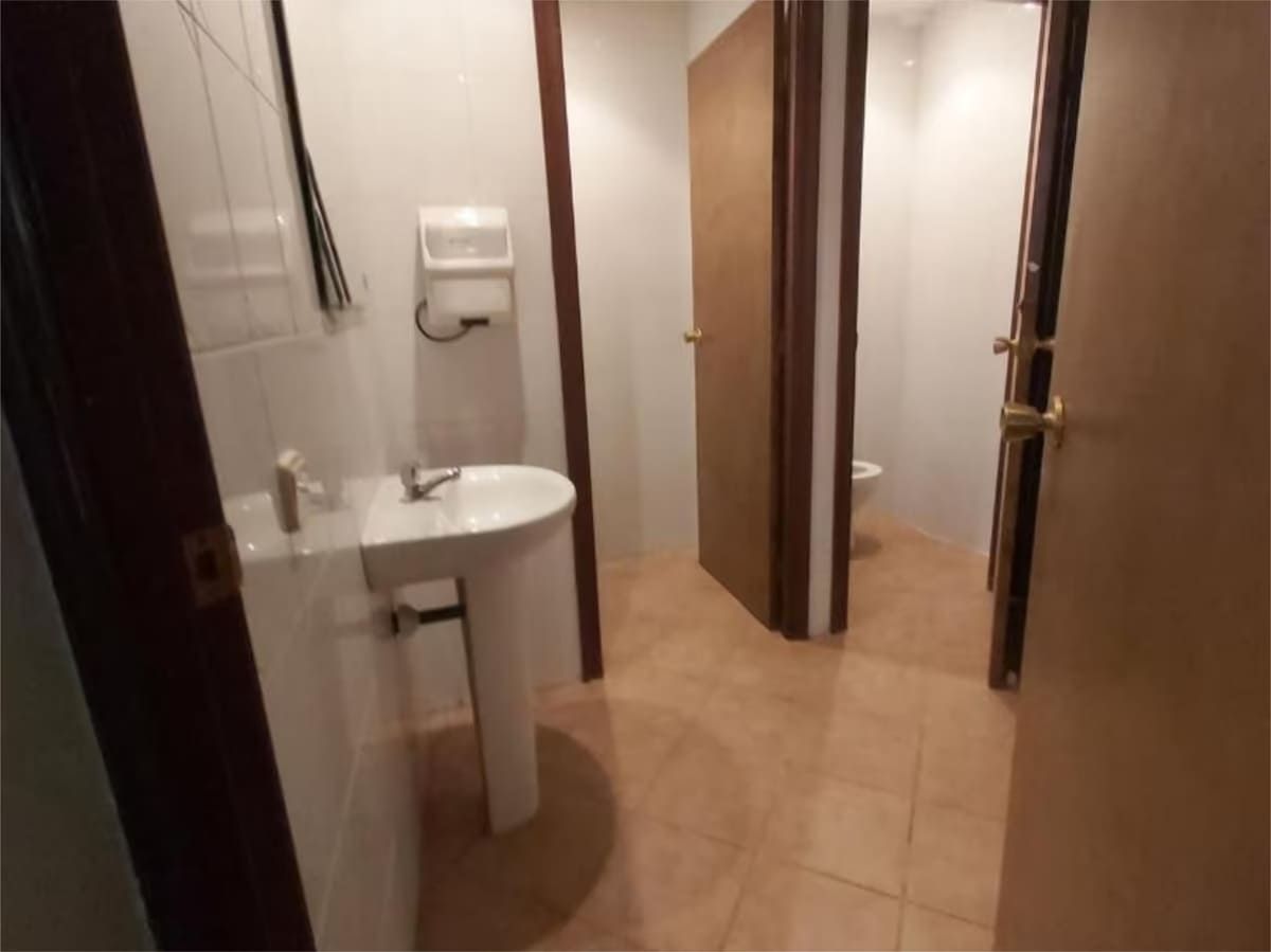 Baño público con lavabo y dos puertas cerradas. Suelo de baldosas beige y paredes claras.