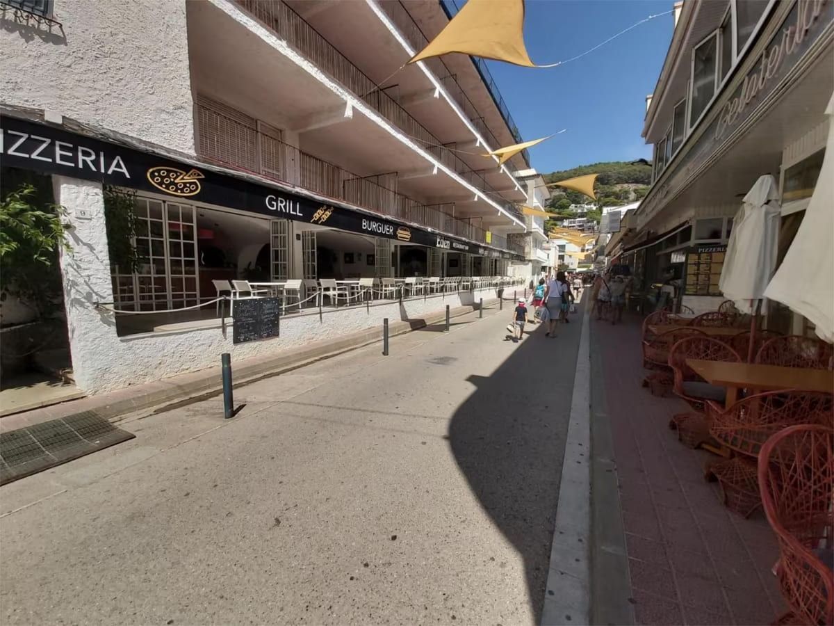 Una calle estrecha llena de tiendas y asientos al aire libre; gente caminando en un día soleado.