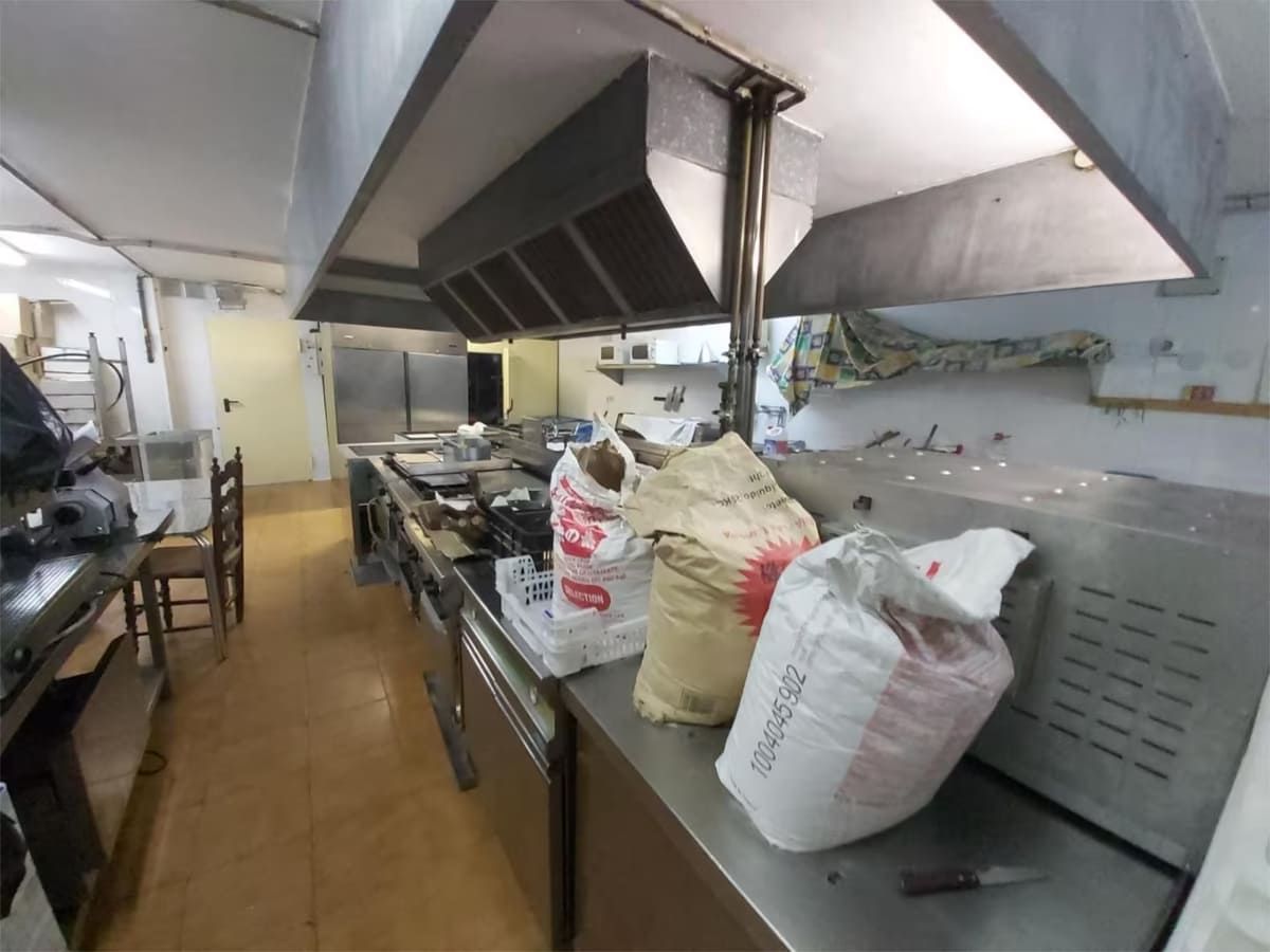 Una cocina comercial con encimeras y electrodomésticos de acero inoxidable, incluyendo una estufa y una campana extractora. Hay bolsas con suministros sobre la encimera.