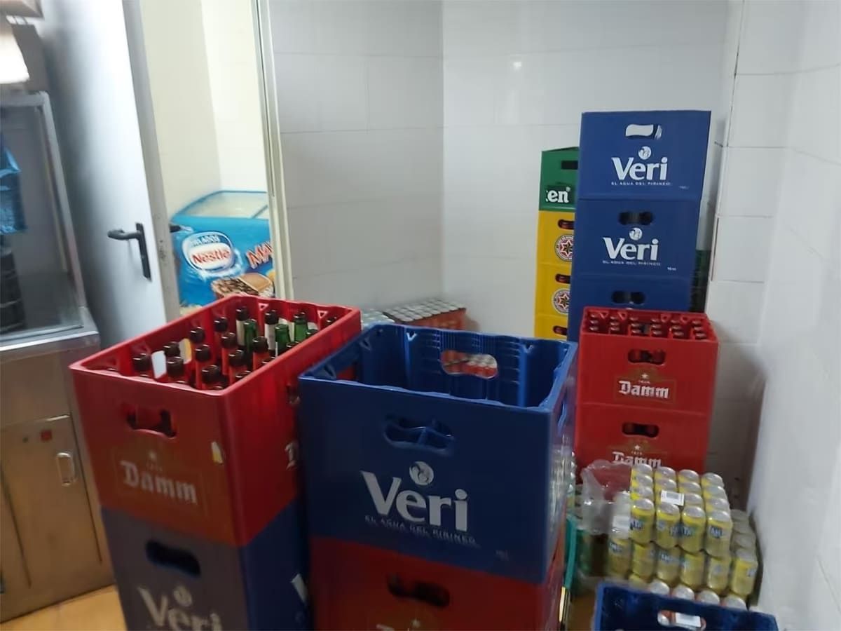 Cajas de cerveza y refrescos apiladas en una habitación con un congelador y una puerta visibles.