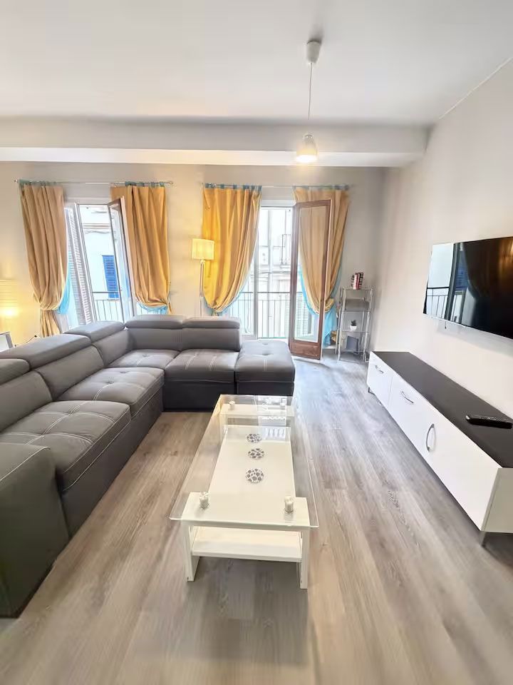 Sala de estar moderna y luminosa con sofá modular gris, mesa de centro blanca, televisor de pared y cortinas amarillas.