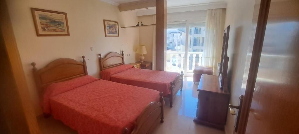 Habitación pequeña con dos camas rojas, armario y ventana con cortinas claras.