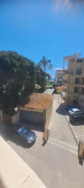 Calle soleada con coches aparcados, edificios de apartamentos y árboles junto a una carretera estrecha.