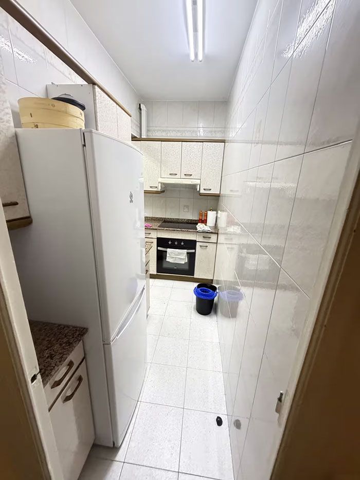 Cocina estrecha y blanca tipo galera con refrigerador, armarios, estufa y un cubo de basura azul en el suelo.