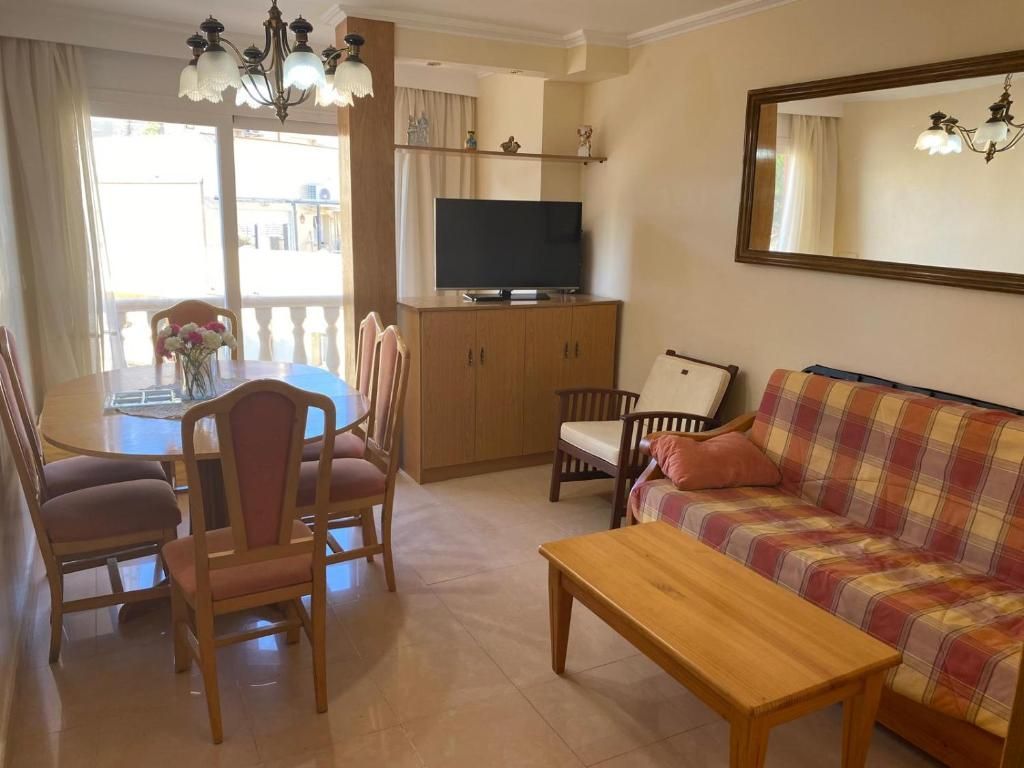 Sala de estar luminosa con sofá a cuadros, mesa de comedor de madera, mueble para la televisión, espejo y lámpara de araña cerca de una ventana.