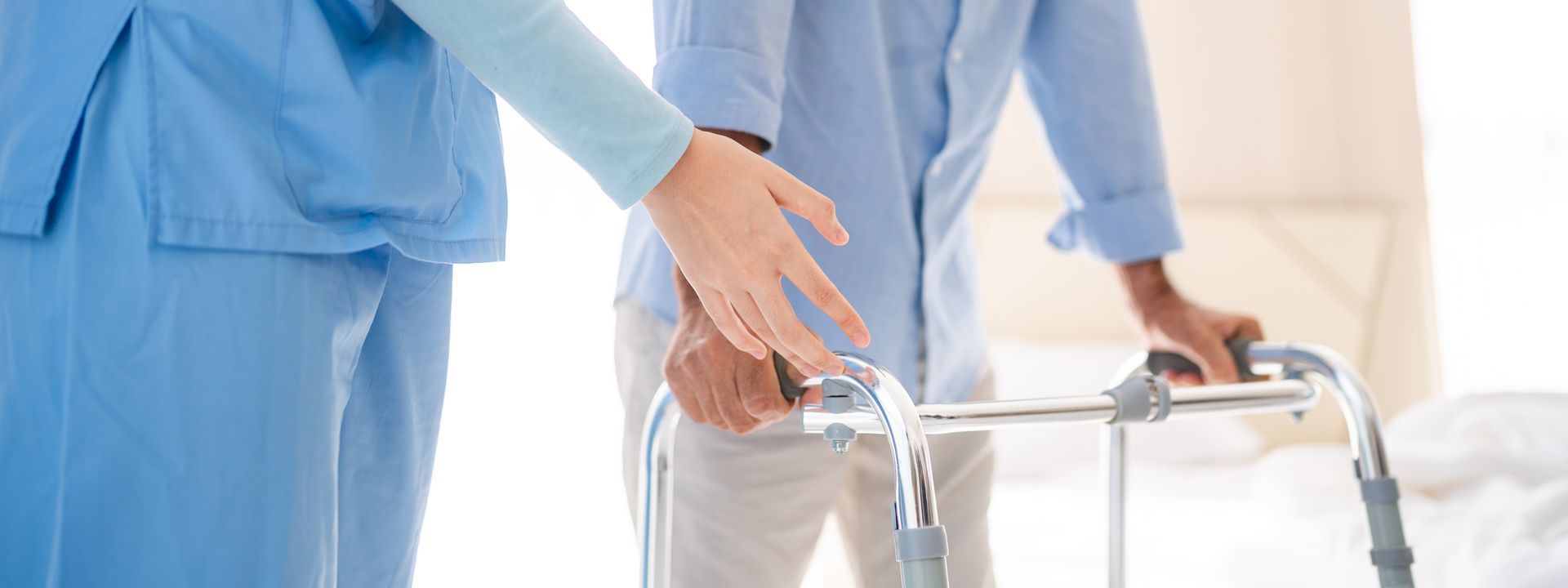 Dans une pièce lumineuse, une infirmière en blouse bleue aide une personne âgée à marcher avec un déambulateur.