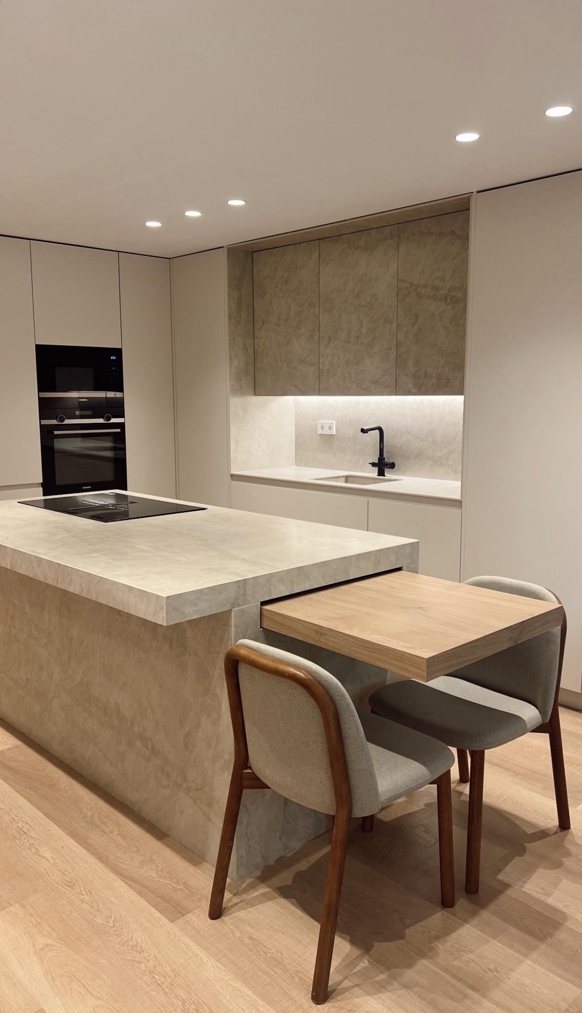 Cocina moderna con isla, horno empotrado y una pequeña mesa de comedor con sillas. Esquema de colores claros.