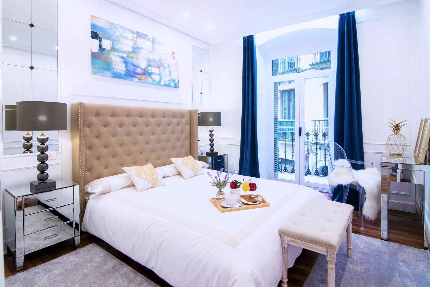 Dormitorio con cama, mesitas de noche con espejo, cortinas azules, obras de arte y vistas desde el balcón.