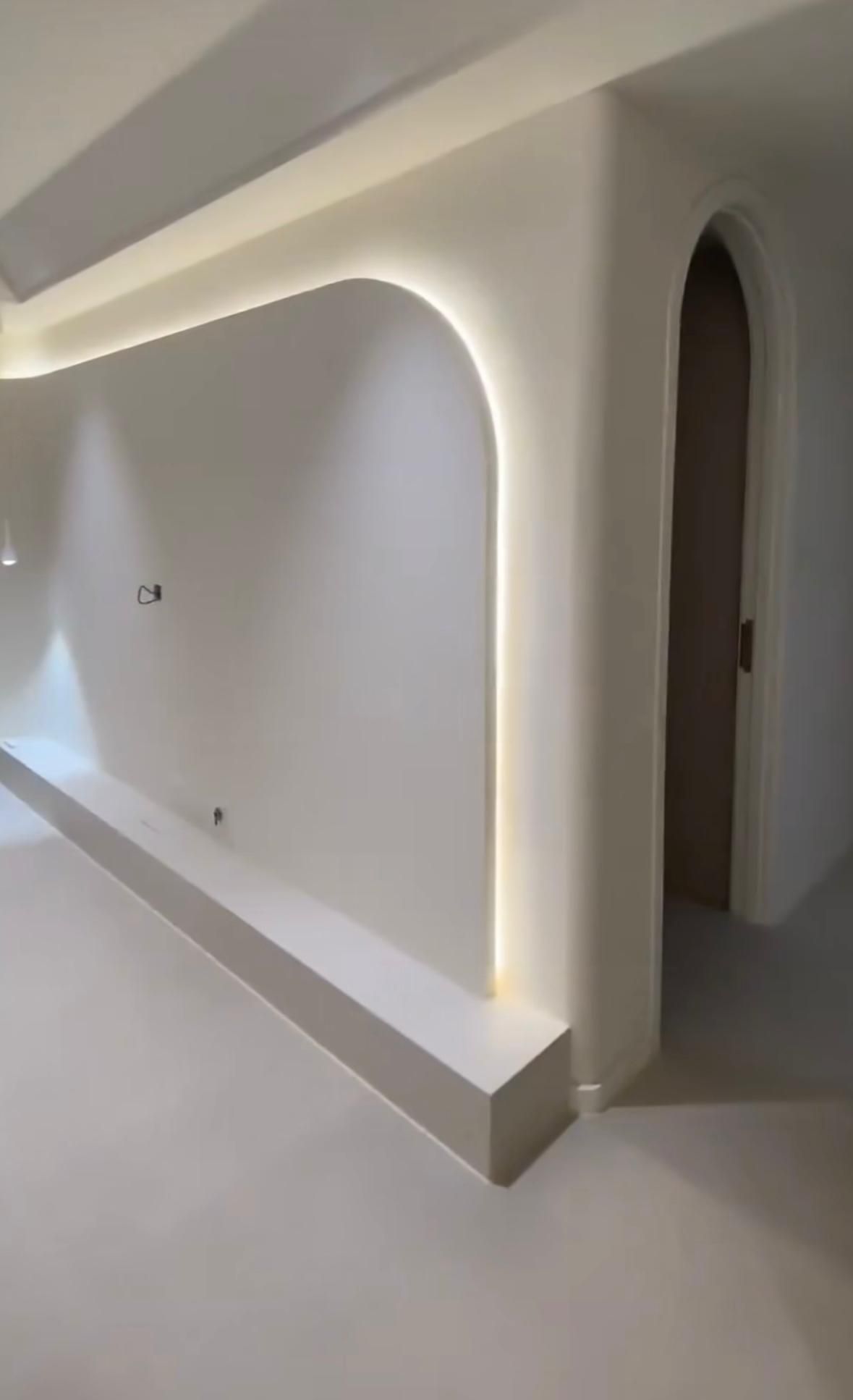 Pared blanca con detalles arqueados e iluminación empotrada, que conduce a una puerta arqueada.