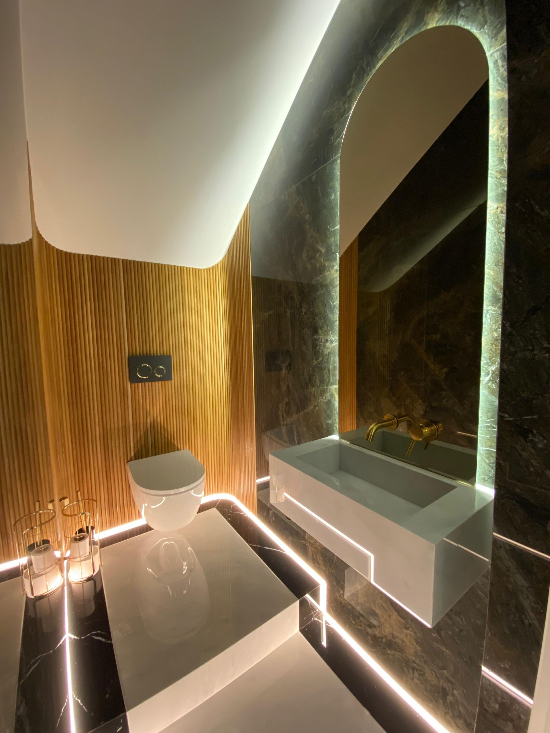 Baño moderno con paneles de madera, inodoro blanco, bañera y suelos de mármol con iluminación LED.