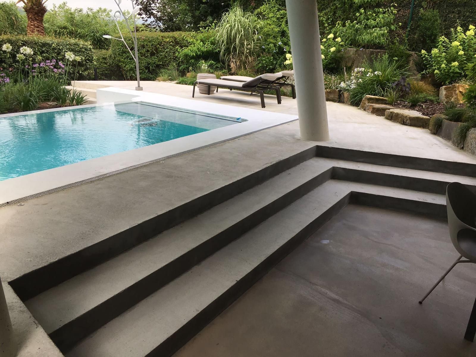 Patio de hormigón con escalones que conducen a la piscina. Una exuberante vegetación rodea el espacio exterior.