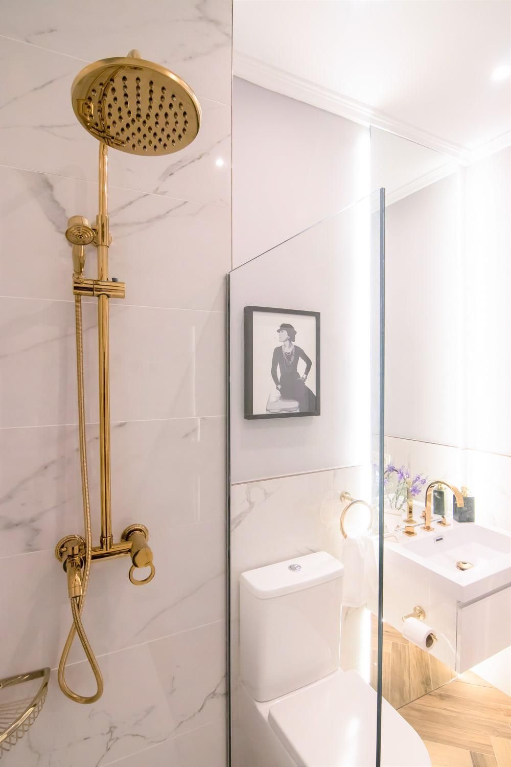 Grifo de ducha dorado en un baño con azulejos de mármol. Un retrato cuelga sobre el inodoro, cerca del lavabo.