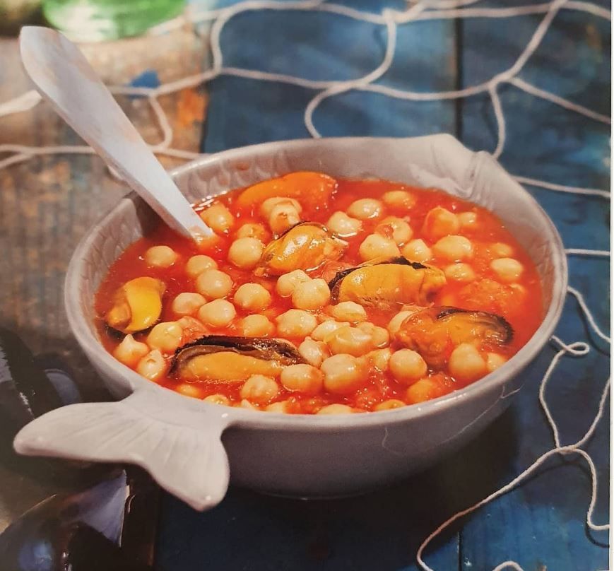 Un tazón de sopa con garbanzos y mejillones con una cuchara dentro.