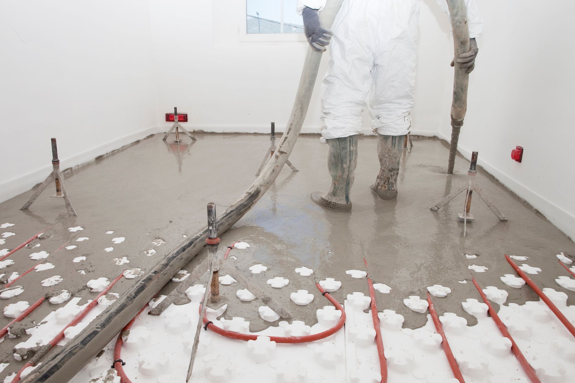 Une personne en combinaison de protection verse du béton sur un plancher chauffant dans une pièce.