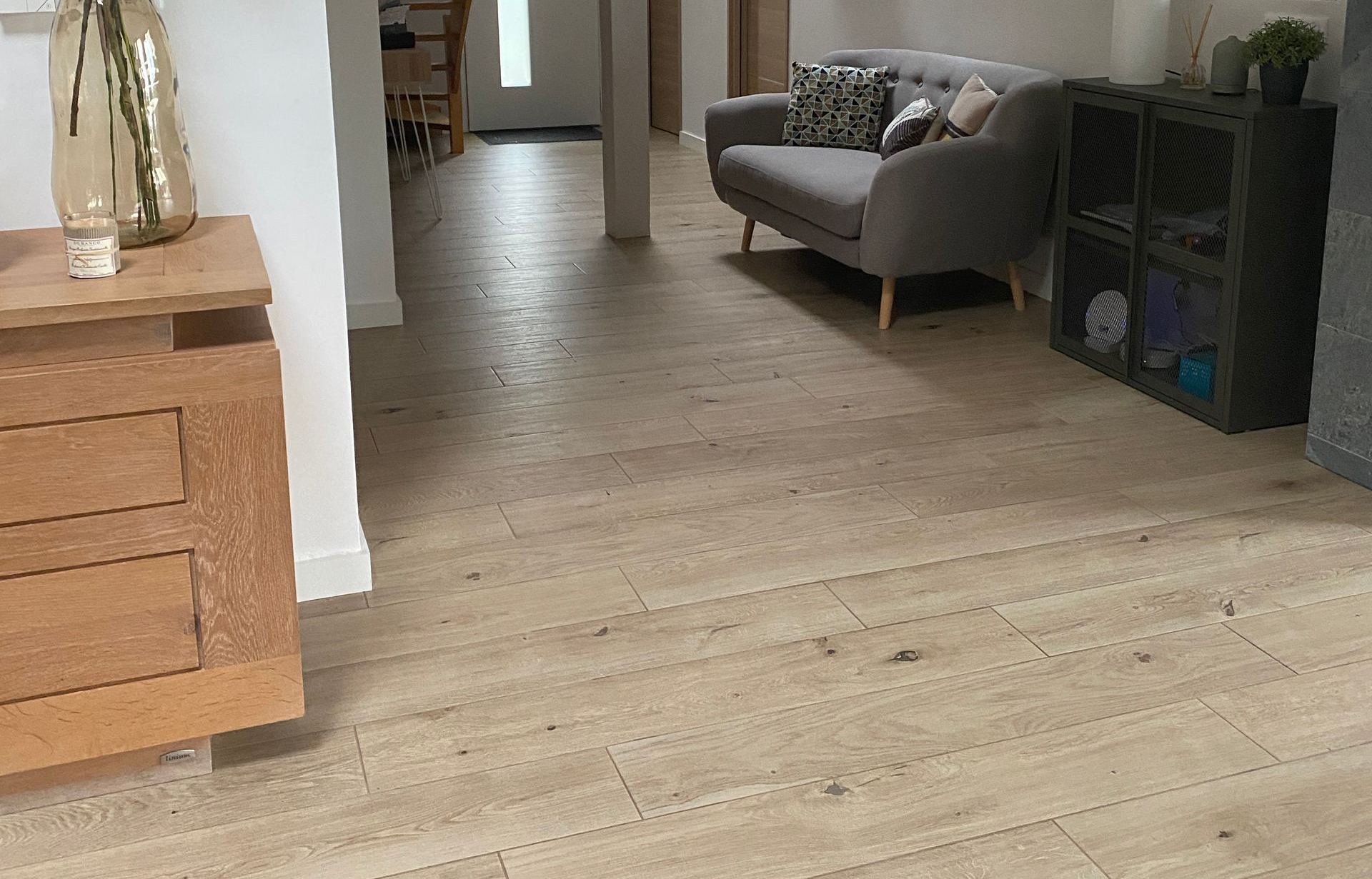 Un parquet en bois clair mène à un salon avec un canapé gris et un meuble.