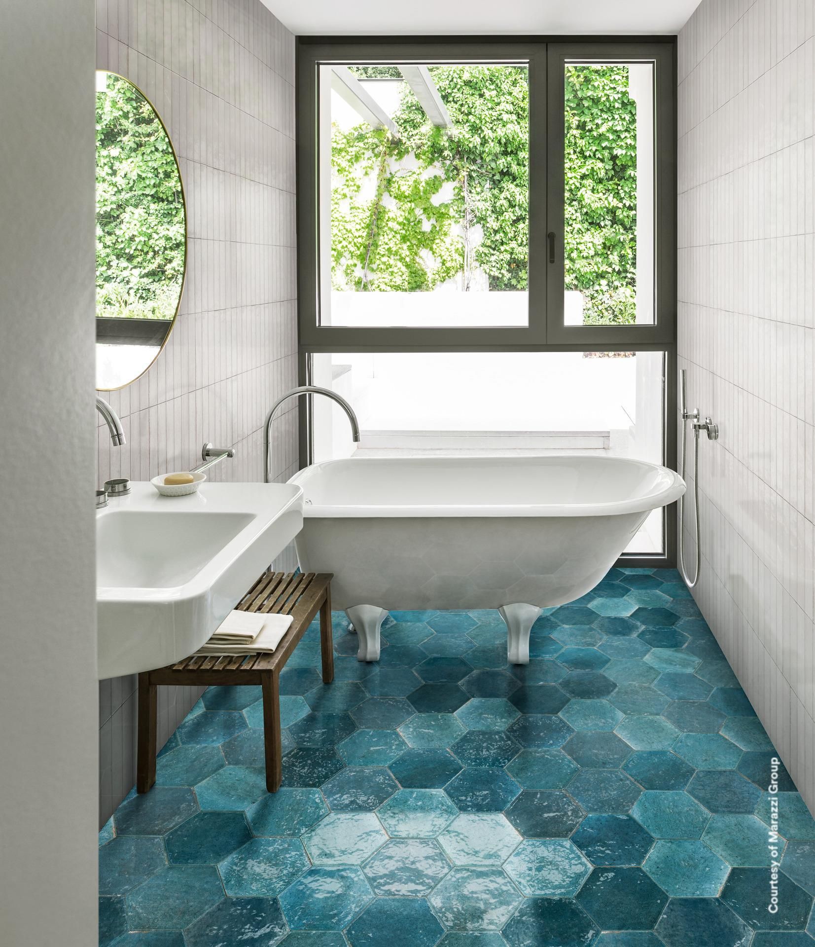 Salle de bain avec sol en carrelage hexagonal bleu, baignoire sur pieds et grande fenêtre donnant sur la verdure.