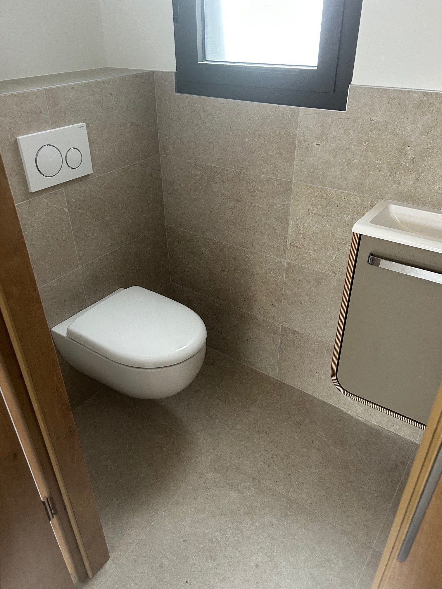 Petite salle de bains avec W.-C suspendu et lavabo, carrelage beige aux murs et au sol, fenêtre, boutons de chasse d'eau.