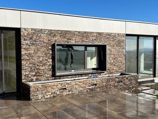 Vue extérieure d'un bâtiment moderne avec mur en pierre et grandes fenêtres sous un ciel bleu.