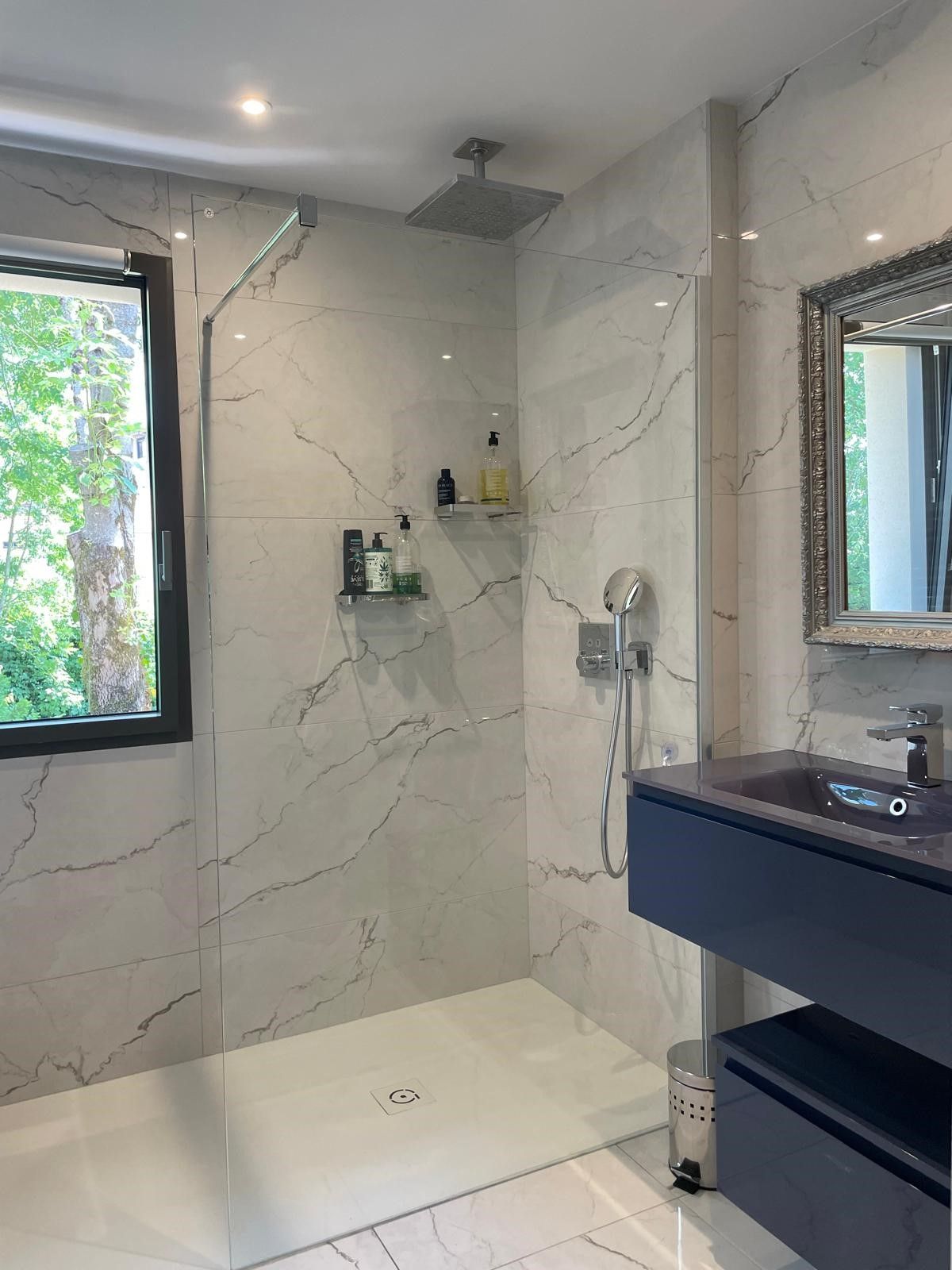 Salle de bains moderne avec carrelage aspect marbre, douche en verre, vanité bleue et grande fenêtre avec vue sur les arbres.