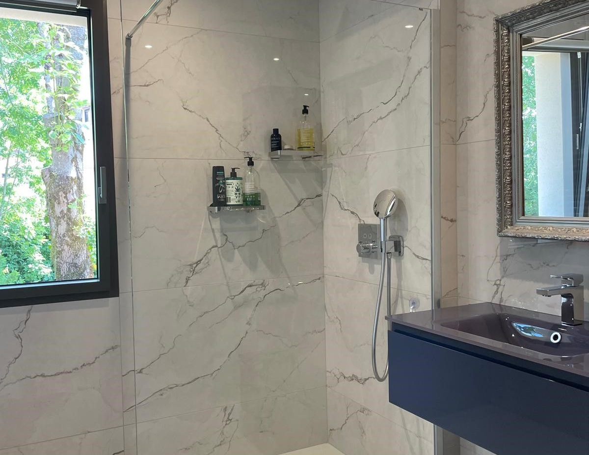 Salle de bains moderne avec carrelage imitation marbre blanc, douche en verre, vanité bleue, fenêtre avec vue sur les arbres.