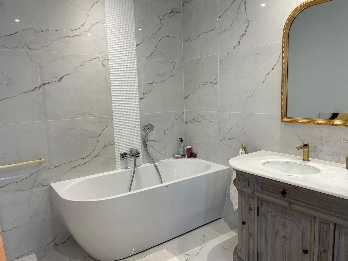 Salle de bains avec baignoire blanche, murs en marbre, vanité en bois et miroir à cadre doré.