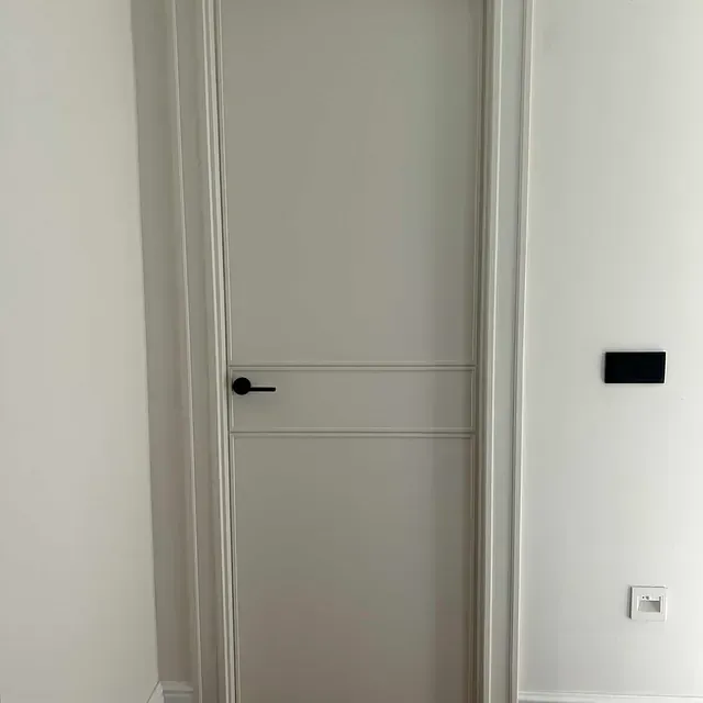 Una puerta blanca con una manija negra está en una habitación al lado de una pared blanca.