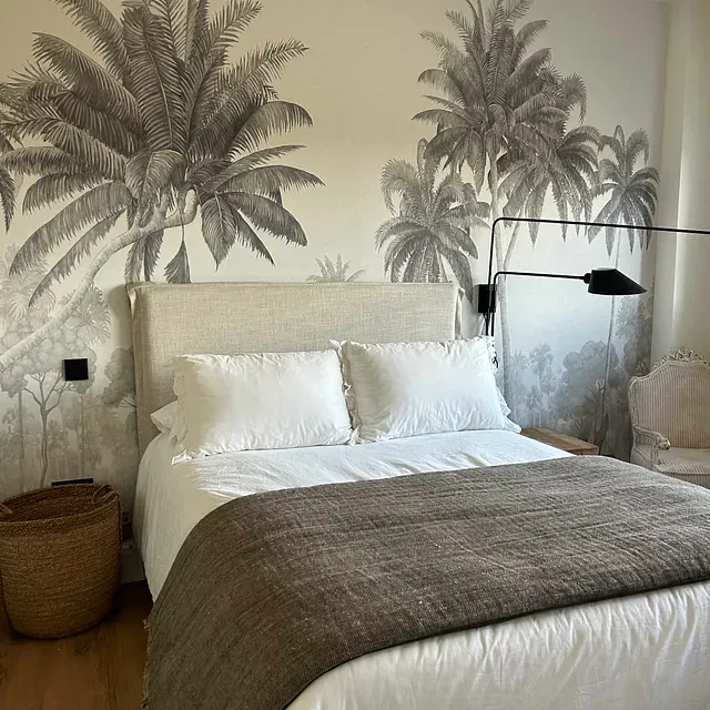 Un dormitorio con una cama, silla, lámpara y palmeras en la pared.