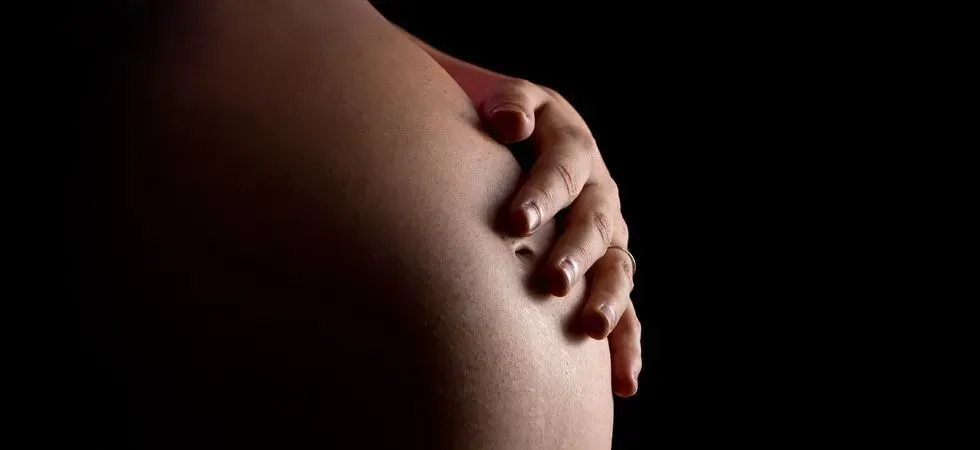 L'homme sourit en écoutant le ventre de la femme enceinte. Il lui caresse l'abdomen ; elle porte un pantalon gris et un haut blanc.