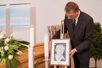 Ein Mann im Anzug hält das Bild einer Frau vor einem Sarg.