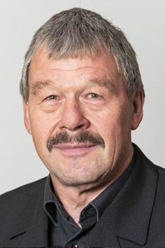 Ein Mann mit Schnurrbart trägt einen Anzug und ein schwarzes Hemd.