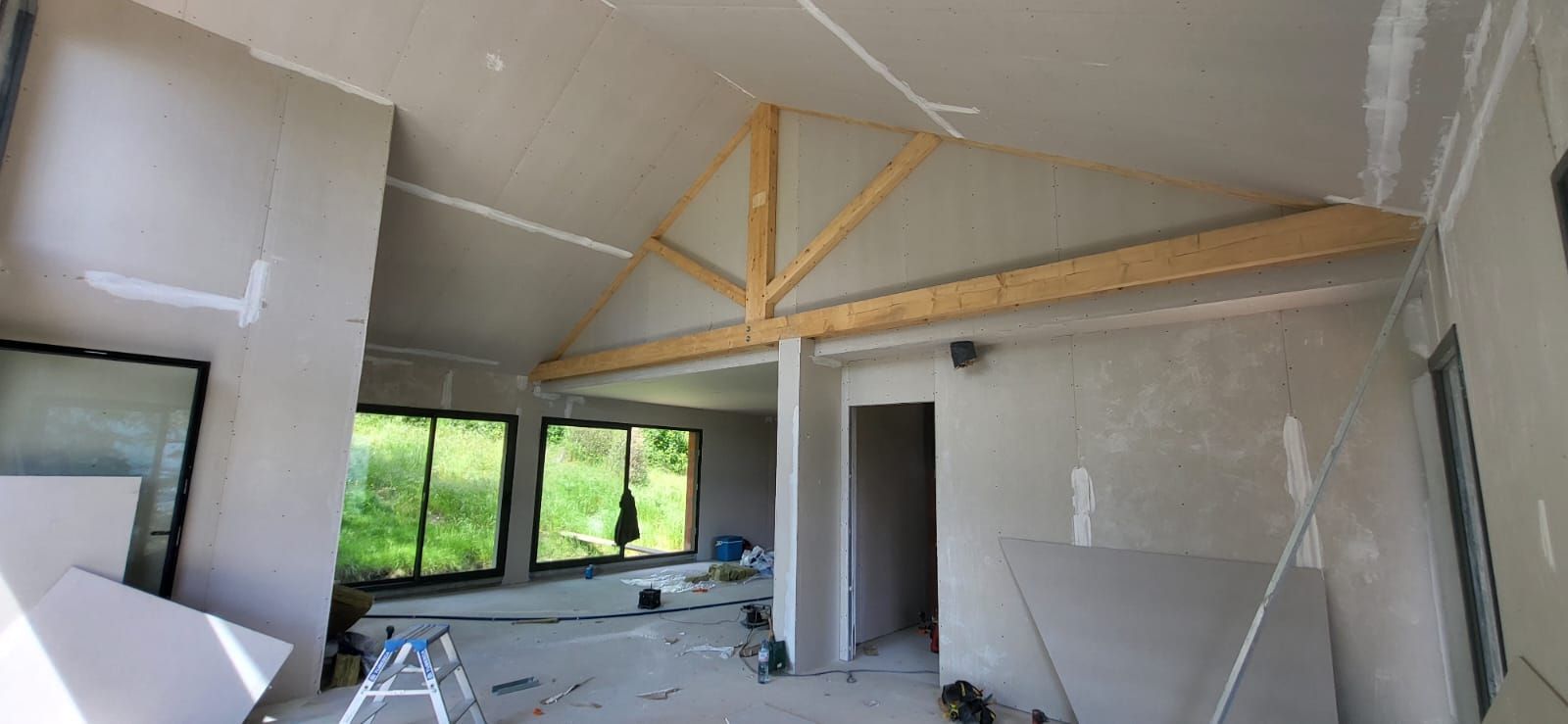 Intérieur d’une pièce en construction avec des poutres apparentes, des murs, des fenêtres et des matériaux de construction au sol.