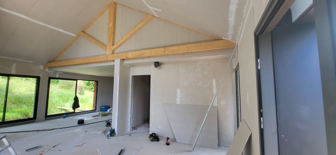 Pièce partiellement terminée, avec des fermes en bois apparentes, des murs, de grandes fenêtres et des matériaux de construction éparpillés à l’intérieur.